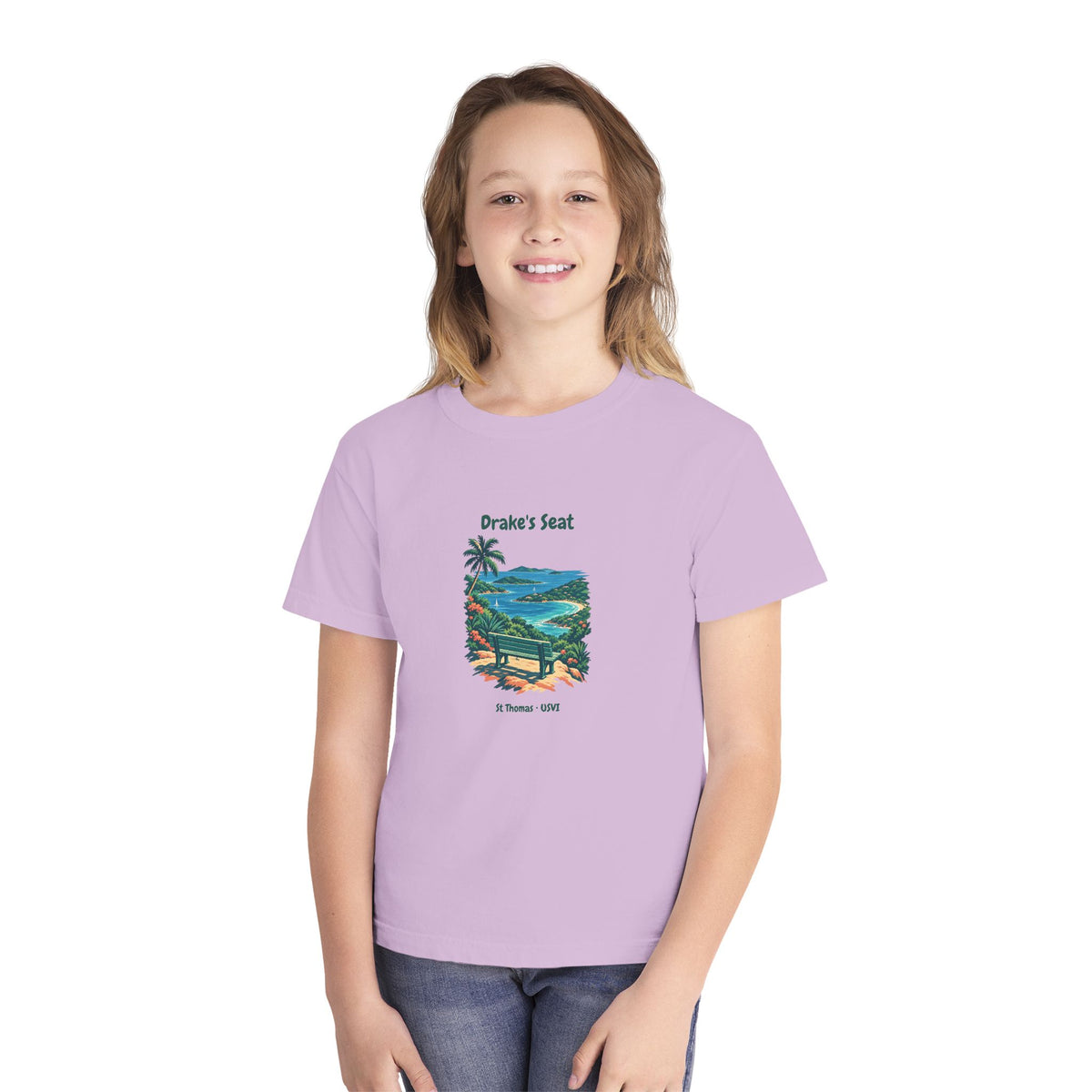 A young girl with light brown hair smiling, wearing a light purple t-shirt with a 'Drake's Seat' beach graphic and blue jeans, set against a plain white background.